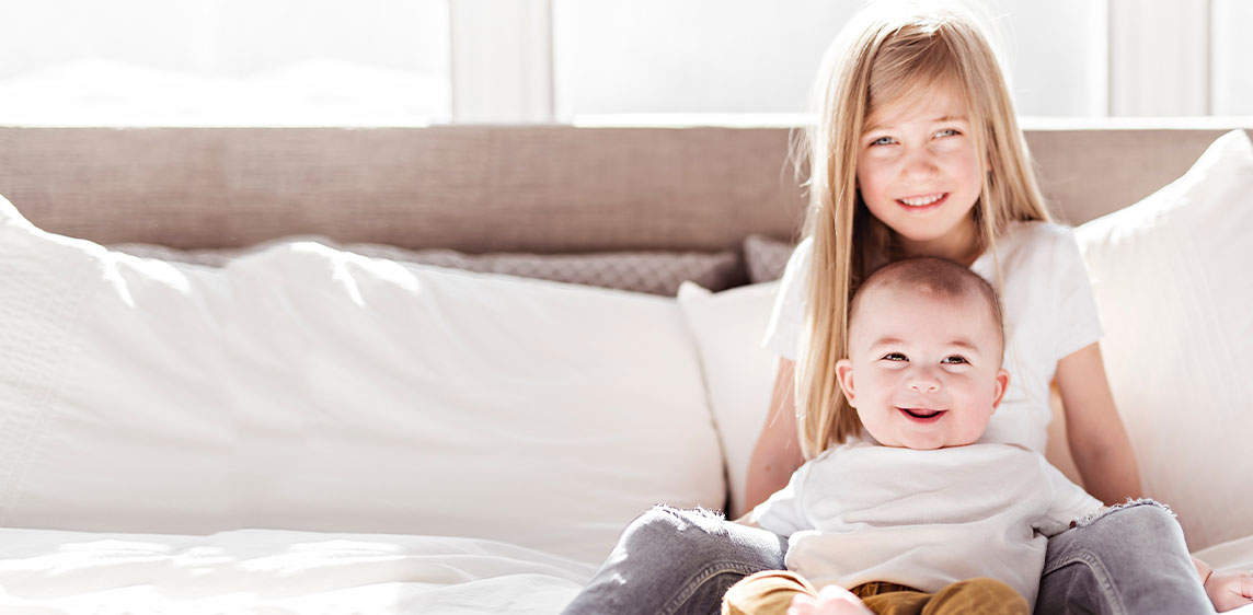 Emsan® Babys und Kinder Ein Mädchen sitzt mit seinem kleinen Geschwisterchen auf einem Bett.