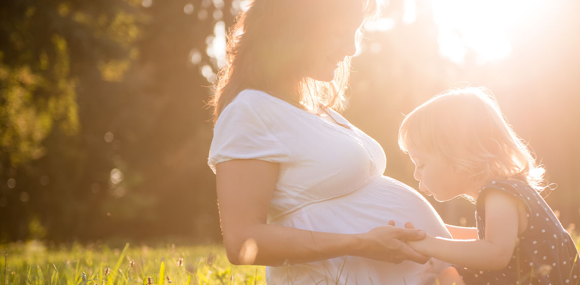 Emsan® Allergieprodukte Eine schwangere Frau sitzt mit ihrem Kleinkind im Gras. Das Kind küsst den Babybauch.