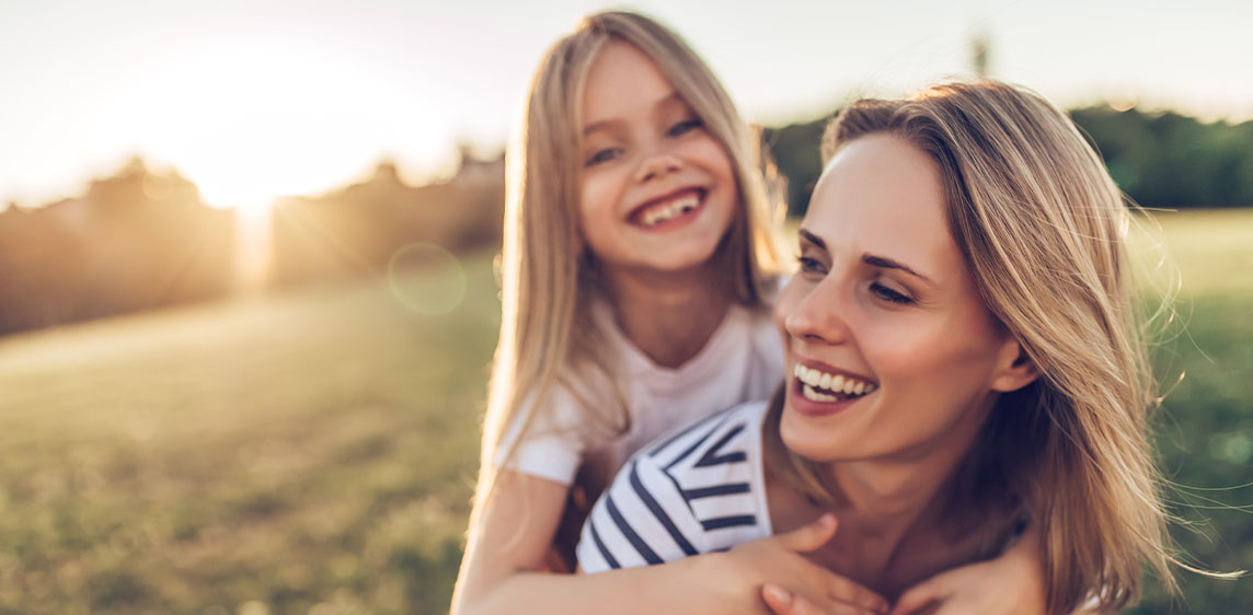 Mutter und Tochter lachen zusammen auf einer Wiese im Licht der untergehenden Sonne.