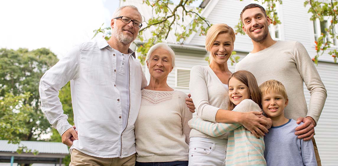 Eine Familie aus drei Generationen steht zusammen vor einem Haus im Garten und lächelt.