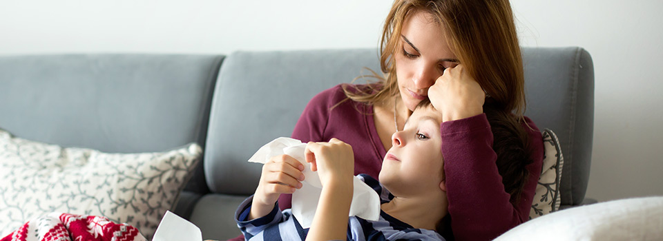 Häufig ist eine Erkältung die Ursache für Halsweh bei Kindern. Eine Mutter tröstet ihr krankes Kind. Beide liegen auf der Couch.