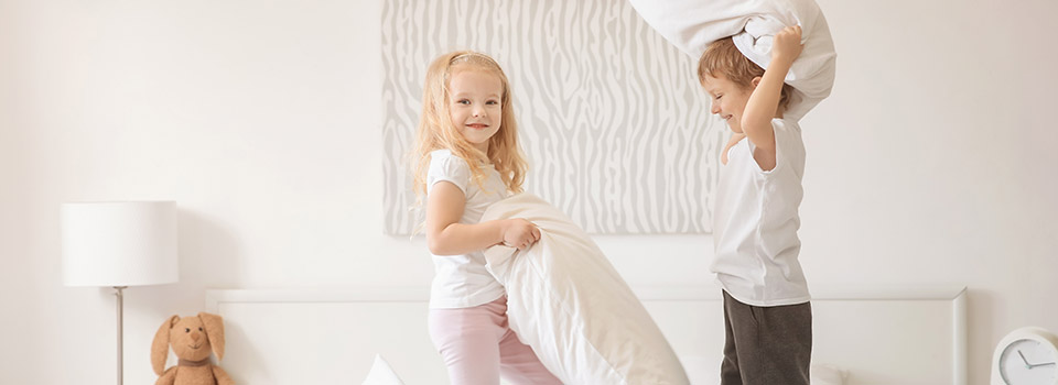 In Kinderbetten finden Hausstaubmilben paradiesische Bedingungen vor. Ein Junge und ein Mädchen stehen auf einem Bett und haben jeweils ein Kissen in der Hand.