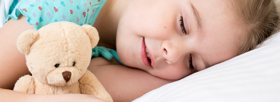 Die Stofftiere des Kindes sollten regelmäßig von Hausstaubmilben befreit werden. Kind liegt mit Teddybär im Arm mit geschlossenen Augen im Bett.