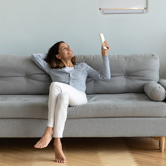 Eine Frau sitzt auf einer Couch und richtet eine Fernbedienung auf die darüber angebrachte Klimaanlage.