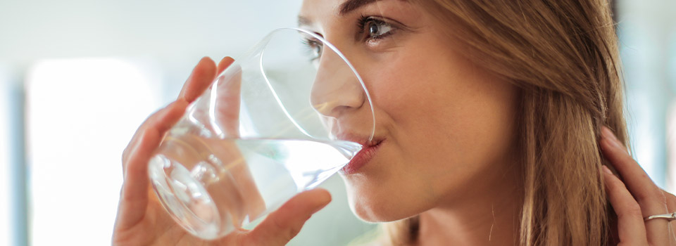 Vor und nach dem Tragen der Atemschutzmaske sollten Sie auf eine ausreichende Flüssigkeitszufuhr achten. Frau trinkt Wasser aus einem Glas