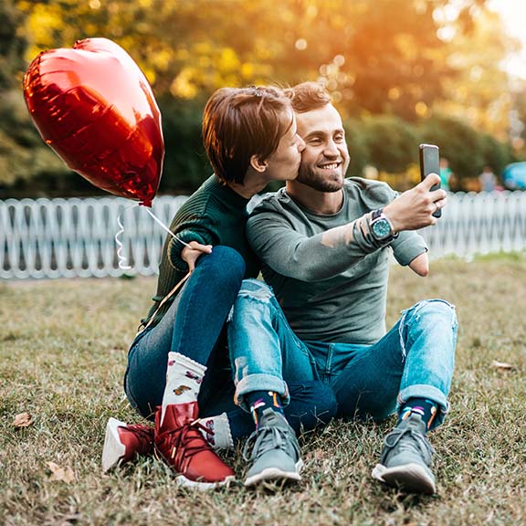 Ein Pärchen sitzt mit Herzluftballon auf einer Wiese und macht ein Selfie. 