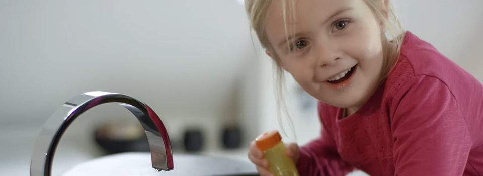Nasenspülungen eignen sich bereits für Kinder ab 3 Jahren – mit Unterstützung der Eltern. Ein lächelndes Mädchen lehnt am Waschbecken und hält die Kindernasendusche in der Hand.