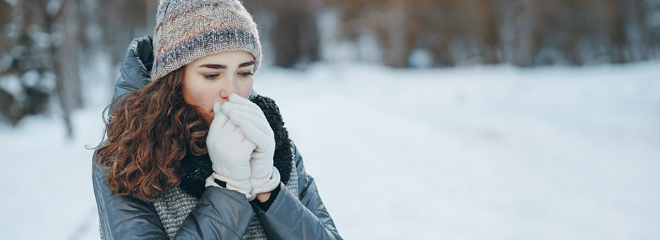Halsschmerzen klingen meist nach wenigen Tagen ab. Eine junge Frau steht im Schnee. Sie trägt Winterkleidung und haucht in ihre Handschuhe.