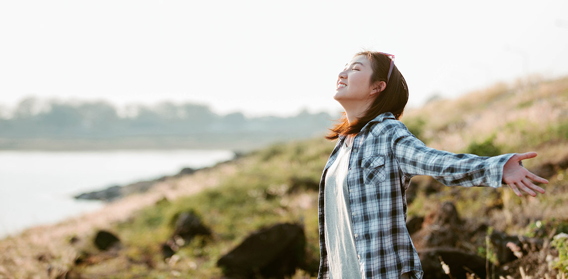 Erkältung – was nun? Eine junge Frau steht in der Natur und atmet tief durch.