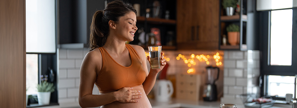 Schwangere sollten täglich 1,5 Liter Flüssigkeit trinken, um die Schleimhäute feucht zu halten. Eine Schwangere steht mit einem Glas Wasser in der Küche. Ihre rechte Hand liegt auf ihrem Bauch.