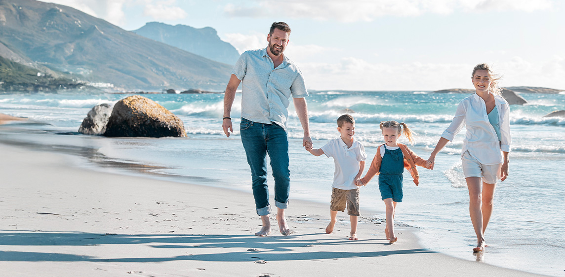 Eine Familie geht in sommerlichem Outfit barfuß am Stand entlang und lächelt in die Kamera
